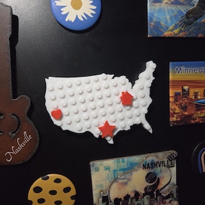 May include: A collection of souvenir magnets on a black surface. The magnets include a white map of the United States with red star and heart accents, a blue daisy, a guitar with "Nashville" text, and a yellow cookie with black spots.