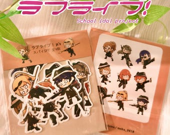 Pode incluir: Um conjunto de nove adesivos com personagens de anime do projeto Love Live! School Idol. Os adesivos são impressos em papel branco e têm um acabamento brilhante. Os personagens usam roupas pretas e brancas e seguram vários adereços, incluindo armas de fogo, espadas e chapéus. O texto nos adesivos diz "Love Live! µ's Spy All 9".