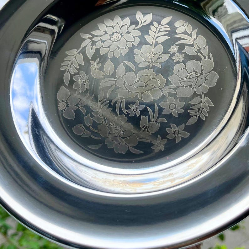 May include: A polished, silver-coloured metal plate with an etched floral pattern in the centre. The design includes various flowers and leaves. The plate has a broad, curved rim and reflects the surrounding environment.