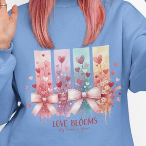 May include: A light blue sweatshirt with a floral and heart design. The design includes the text "LOVE BLOOMS My Heart is Yours". The person in the image is wearing a blue beanie and holding a black smartphone.