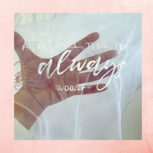 May include: A sheer white veil featuring white embroidered text that reads "AFTER ALL THIS TIME? always" followed by the date "29/08/2026" in a script font. The fabric is translucent, allowing the texture and colour of the surface behind it to show.