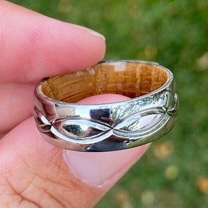 May include: A silver ring with a wood inlay and a wave-like design. The ring is on a finger.