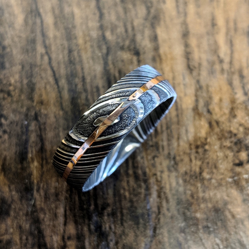 May include: A silver and rose gold wedding band with a patterned design. The band is made of metal and has a polished finish.