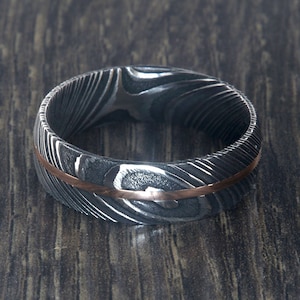 May include: A silver and rose gold wedding band with a patterned design. The band is made of Damascus steel and has a thin rose gold inlay.