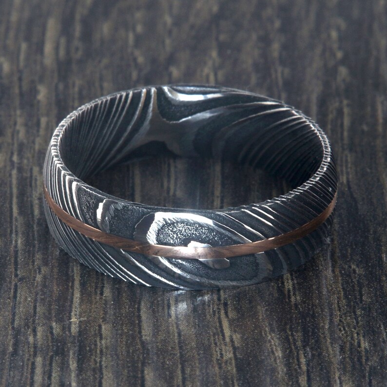 May include: A silver and rose gold wedding band with a patterned design. The band is made of Damascus steel and has a thin rose gold inlay.