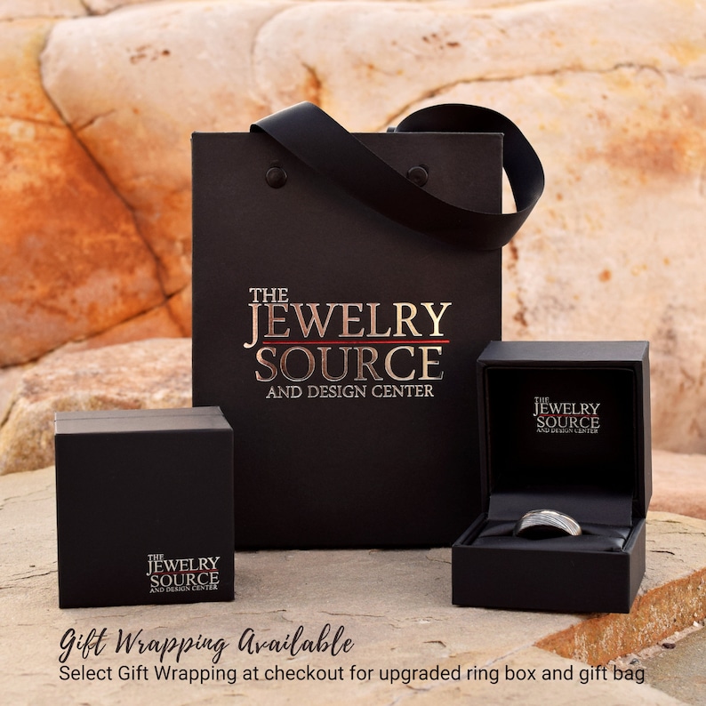 May include: A black gift bag with the text "The Jewelry Source and Design Center" printed on the front. The bag is next to a black ring box with the same text printed on the lid. The ring box is open and a silver ring is inside.