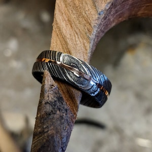 May include: A black and silver ring with a thin gold band. The ring has a textured, patterned surface.