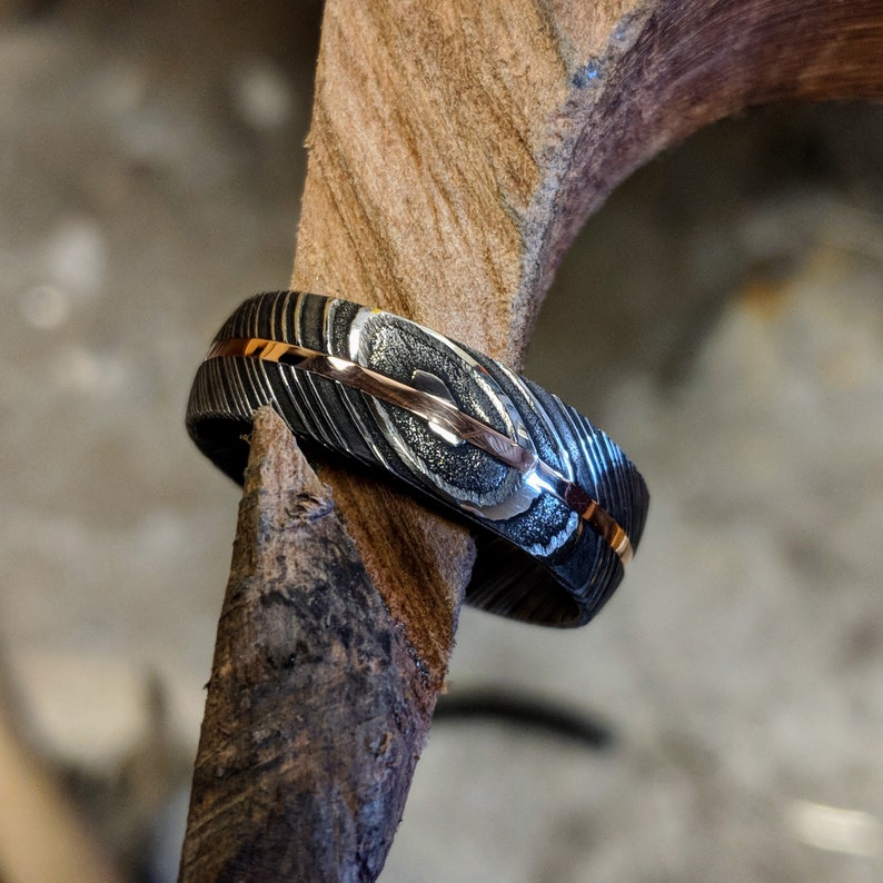 May include: A black and silver ring with a thin gold band. The ring has a textured, patterned surface.