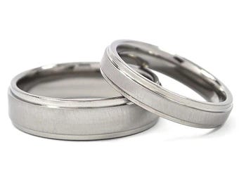 May include: Two silver wedding bands with a brushed finish. The band on the left has a beveled edge, while the band on the right has a smooth edge.