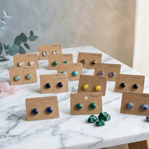 Könnte beinhalten: Eine Sammlung von Ohrsteckern aus Edelsteinen, präsentiert auf kleinen braunen Karten. Die Ohrringe zeigen verschiedene Steine, darunter Rosenquarz, Amethyst und Malachit. Sie sind auf einer weißen Marmoroberfläche angeordnet.