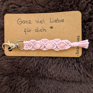 Könnte beinhalten: Ein rosa Makramee-Schlüsselanhänger mit drei rosa Perlen und einem silbernen Verschluss. Der Schlüsselanhänger ist an einer braunen Karte mit dem deutschen Text "Ganz viel Liebe für dich" und einem Herzsymbol befestigt.