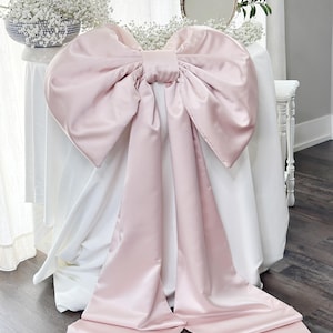 May include: A large, light pink satin bow draped over a white tablecloth. The bow has long, flowing tails. The background includes a white table with a blue and white bowl of flowers, a mirror, and greenery.