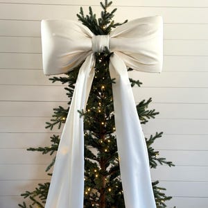 May include: A large, ivory-colored bow sits atop a Christmas tree. The bow is made of a soft, flowing fabric and has long, draping tails. The tree is decorated with small, warm white lights, creating a festive holiday display.