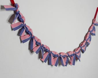May include: A red, white, and blue ribbon garland with bows tied at regular intervals. The garland is hung on a white wall.