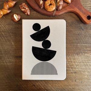 May include: A beige notebook with a black geometric design featuring stacked circles and half circles. The design includes a striped half circle at the bottom.