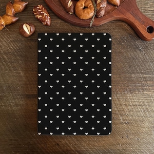 Könnte beinhalten: Ein schwarzes Notizbuch mit einem Muster aus kleinen weißen Herzen. Das rechteckige Notizbuch liegt auf einer Holzoberfläche. Gebäck, darunter Brezeln und Bagels, sind darum herum angeordnet.