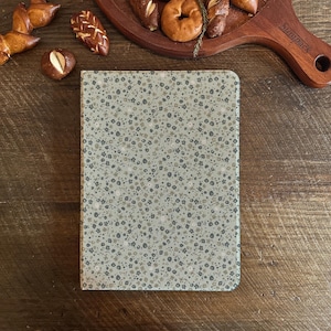 Könnte beinhalten: Ein Notizbuch mit floralem Muster, mit hellgrünem Hintergrund und kleinen grauen und braunen Blumen. Das Notizbuch liegt auf einer Holzoberfläche, im Hintergrund befinden sich Backwaren wie Brezeln und Bagels.