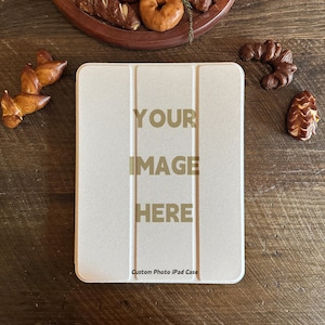Könnte beinhalten: Eine cremefarbene iPad-Hülle mit dem Text "YOUR IMAGE HERE" in goldfarbenen Buchstaben. Die Hülle hat eine strukturierte Oberfläche und ein dreifaches Design. Die Worte "Custom Photo iPad Case" sind unten aufgedruckt. Gebäck ist um die Hülle herum auf einer Holzoberfläche angeordnet.