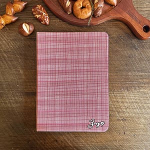 Könnte beinhalten: Ein rosa Notizbuch mit einem Karomuster und dem handgeschriebenen Wort "Zug" mit einem Herzsymbol. Das Notizbuch liegt auf einer Holzoberfläche mit verschiedenen Backwaren, darunter Brezeln und Bagels.