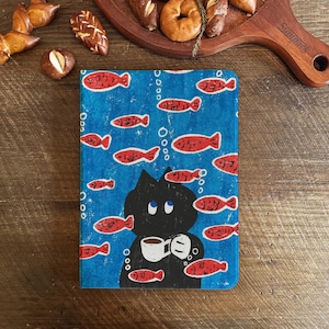 May include: A blue notebook with a quirky design of a black cat holding a coffee cup, with red fish and bubbles. The notebook is rectangular and has a soft cover. The background is a textured blue.