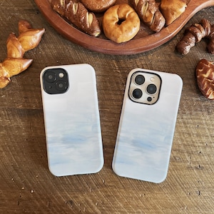 Könnte beinhalten: Zwei weiße Handyhüllen mit blauem Aquarelldesign. Die Hüllen liegen auf einer Holzoberfläche, im Hintergrund ein Holztablett mit Backwaren, darunter Bagels und Brot.