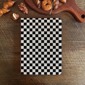 May include: A black and white checkered notebook on a wooden surface. The notebook's cover features a classic checkerboard pattern. Baked goods, including pretzels and bagels, are arranged on a wooden serving board in the background.