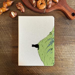 May include: A notebook with a cream-colored cover featuring a black cat silhouette perched on a green illustrated cliff. The notebook is rectangular and has a simple design. Baked goods are in the background.
