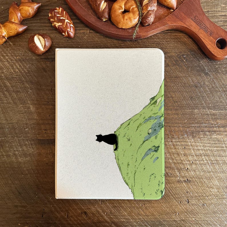 May include: A notebook with a cream-colored cover featuring a black cat silhouette perched on a green illustrated cliff. The notebook is rectangular and has a simple design. Baked goods are in the background.
