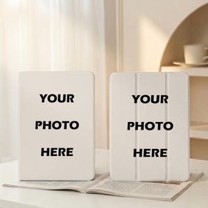 Könnte beinhalten: Zwei cremefarbene Tablet-Hüllen mit dem schwarzen Text "YOUR PHOTO HERE". Eine Hülle ist offen, die andere geschlossen. Sie liegen auf einer weißen Oberfläche, mit einem offenen Buch im Vordergrund.