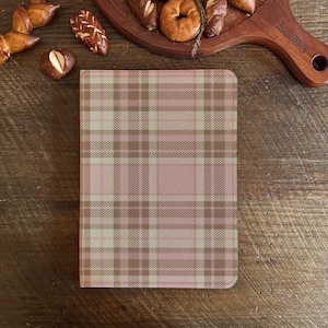 Könnte beinhalten: Ein Notizbuchumschlag mit einem rosa, beigen und braunen Karomuster. Der Umschlag ist rechteckig mit abgerundeten Ecken. Das Notizbuch liegt auf einer Holzoberfläche, im Hintergrund Gebäck.