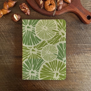 Könnte beinhalten: Ein rechteckiges Notizbuch mit einem Limettenscheibenmuster in Grün- und Weißtönen. Das Notizbuch liegt auf einer Holzoberfläche, im Hintergrund sind Backwaren zu sehen.
