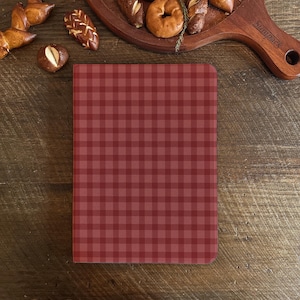 May include: A red and maroon checked notebook on a wooden surface, with baked goods on a wooden serving board in the background. The notebook's pattern is a classic gingham design, ideal for writing or sketching.