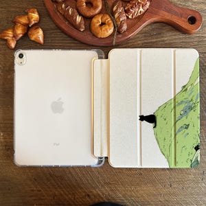 May include: An open iPad with a clear case next to a light-colored case with a green mountain design and a black cat silhouette. The iPad is on a wooden surface with baked goods.