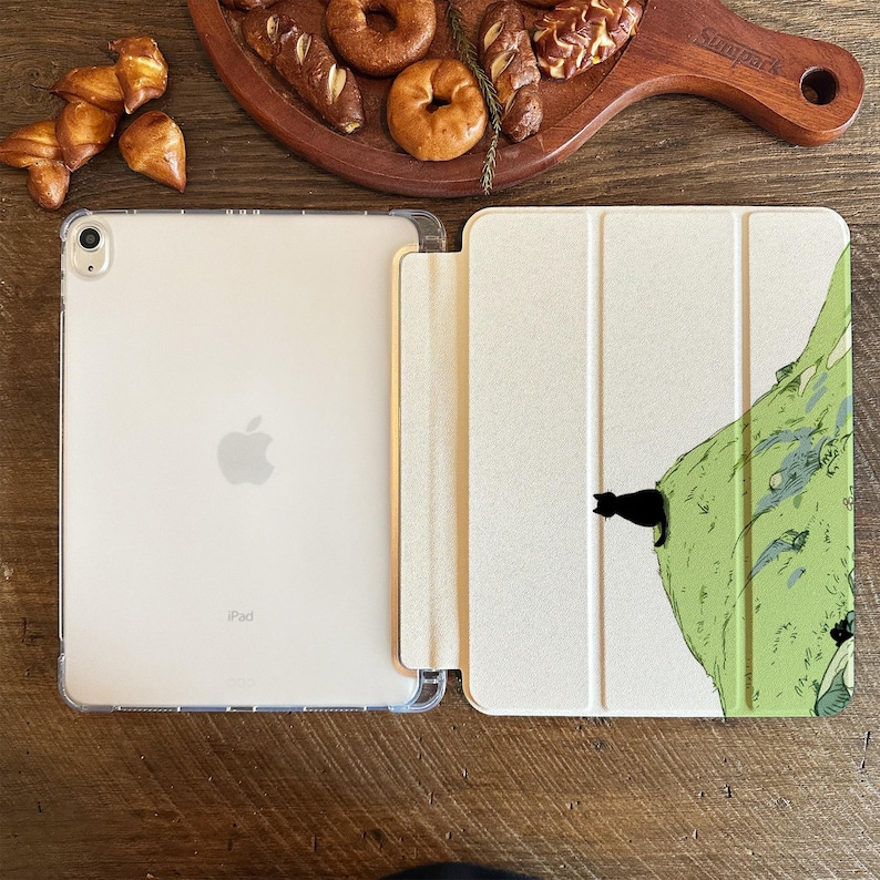 May include: An open iPad with a clear case next to a light-colored case with a green mountain design and a black cat silhouette. The iPad is on a wooden surface with baked goods.