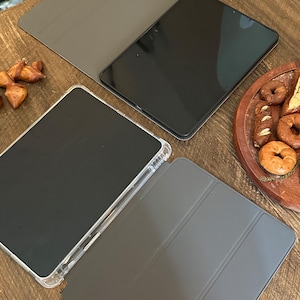 May include: A top-down view showcases two black tablets with protective covers. One tablet is in a clear case, and the other is in a gray cover. The tablets are on a wooden surface, with a tray of baked goods in the background.