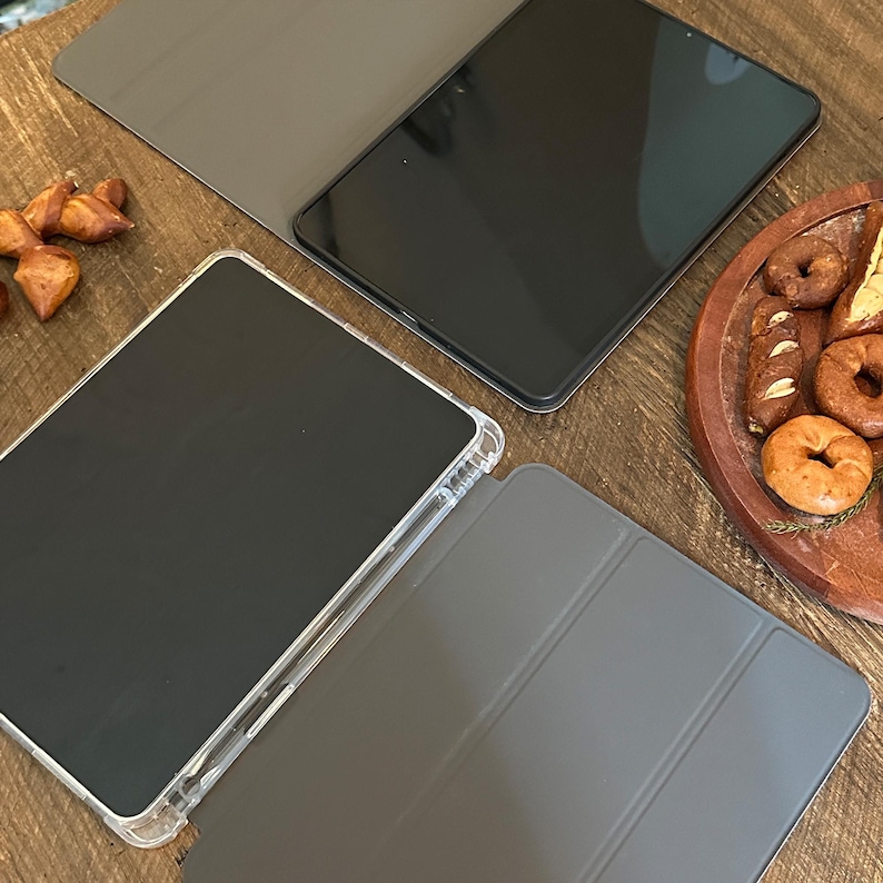 May include: A top-down view showcases two black tablets with protective covers. One tablet is in a clear case, and the other is in a gray cover. The tablets are on a wooden surface, with a tray of baked goods in the background.