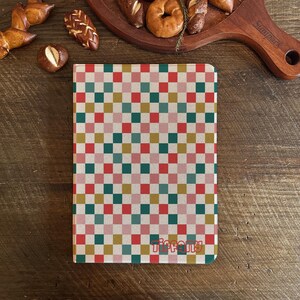 Peut inclure: Un carnet avec un motif à carreaux coloré en carrés rouges, verts, roses et dorés. Le mot "Tiffany" est imprimé en cursive en bas. Le carnet est posé sur une surface en bois avec des pâtisseries en arrière-plan.