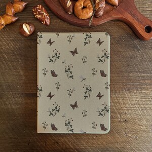 Puede incluir: Una funda beige para tableta con un patrón repetido de mariposas, libélulas y ramitas florales en tonos marrones y verdes. La funda rectangular tiene esquinas redondeadas y está sobre una superficie de madera.