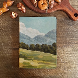 Puede incluir: Una tabla de cortar de vidrio con una escena de paisaje pintada. La escena presenta un cielo azul con nubes blancas, árboles verdes y un campo de hierba en primer plano. Las montañas en el fondo están pintadas en tonos de azul y morado.