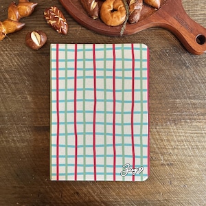 Könnte beinhalten: Ein Notizbuch mit cremefarbenem Einband mit einem Gittermuster aus roten und türkisfarbenen Linien. Das Wort "Zuzg" ist in Kursivschrift unten rechts geschrieben. Gebäck ist um das Notizbuch herum auf einer Holzoberfläche angeordnet.