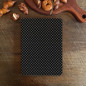 Pode incluir: Um caderno preto com um padrão de bolinhas brancas. O caderno é retangular e está sobre uma superfície de madeira. Vários produtos de panificação, incluindo pretzels e bagels, estão dispostos ao redor do caderno.