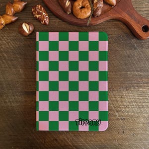 May include: A notebook featuring a pink and green chequered design. The name "Tiffany" is printed in a script font at the bottom. The notebook is on a wooden surface with baked goods in the background.