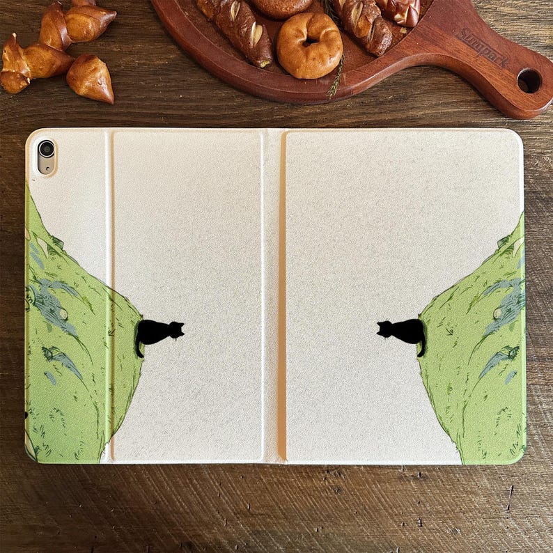May include: An off-white tablet case featuring a whimsical design of a black cat peeking out from a green mountain illustration. The case is open, revealing the tablet screen. The background is a wooden surface.