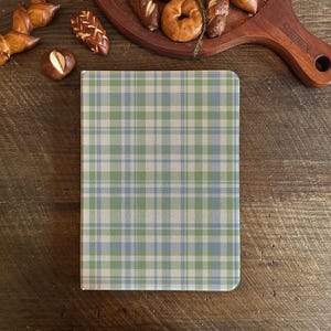 May include: A notebook with a green, blue, and beige plaid pattern. The notebook is on a wooden surface, with baked goods on a wooden serving board in the background.