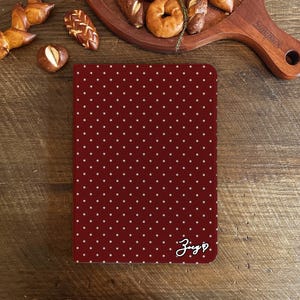 Può includere: Un taccuino bordeaux con un motivo a pois bianchi. Il taccuino ha angoli arrotondati e una firma manoscritta con un simbolo a cuore. I prodotti da forno sono disposti attorno al taccuino su una superficie di legno.