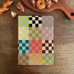 May include: A notebook featuring a vibrant checkerboard design in green, pink, yellow, and black. The notebook is placed on a wooden surface, with baked goods visible in the background.