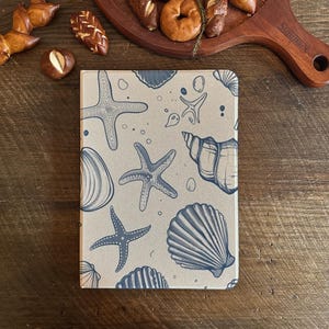 Puede incluir: Una funda beige para tableta con un diseño de conchas marinas y estrellas de mar azules y blancas. La funda rectangular tiene una superficie lisa. El diseño incluye conchas marinas y estrellas de mar de varios tamaños, creando un tema costero.