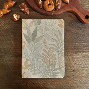 Pode incluir: Um caderno retangular com um padrão de folhas botânicas em tons de verde, bege e dourado. O caderno está sobre uma superfície de madeira, com produtos de panificação ao fundo.
