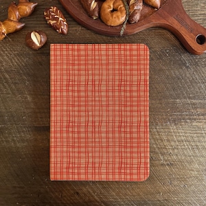 May include: A rectangular notebook featuring a red plaid design on a tan backdrop. The notebook rests on a wooden surface, accompanied by an assortment of baked goods, such as pretzels and bagels, presented on a wooden board.