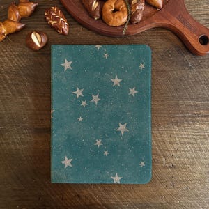 Könnte beinhalten: Ein teal-farbenes Notizbuch mit einem Muster aus weißen Sternen und Sprenkeln. Das rechteckige Notizbuch liegt auf einer Holzoberfläche. Backwaren sind im Hintergrund zu sehen.